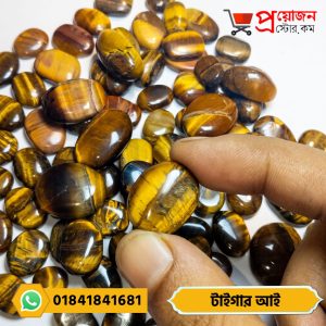 Fingertips holding a smooth golden-yellow natural Tiger's Eye stone cabochon over a pile of gemstones by Proyojonstore