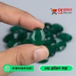 Close-up of a faceted oval vivid green natural Old Brazil Emerald gemstone held between fingertips over loose gems by Proyojonstore